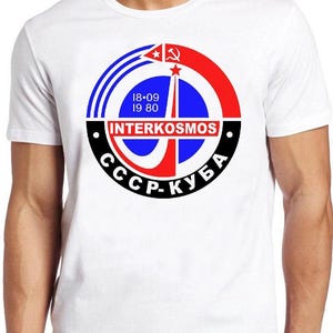 May include: A white short-sleeved t-shirt featuring a circular graphic on the chest. The design includes a red, blue, and black emblem with the text INTERKOSMOS, 18-09 19 80, and CCCP-КУБА, along with a hammer and sickle symbol and a rocket icon.