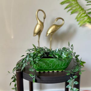 Peut inclure: Un pot de fleurs en céramique verte avec un motif abstrait blanc, rempli de plantes vertes retombantes, est posé sur une table marron foncé. Deux figurines de grues dorées se dressent en arrière-plan.