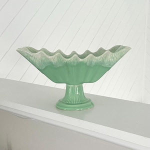 May include: A mint green ceramic centerpiece bowl with a scalloped rim and a pedestal base. The bowl has a textured surface and a light green glaze with a white trim. The bowl is a decorative item.