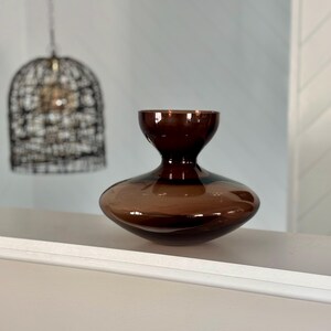 May include: A brown glass vase with a wide, rounded base and a narrow neck. The vase has a dark brown colour and a smooth, glossy finish. The vase is sitting on a white surface.