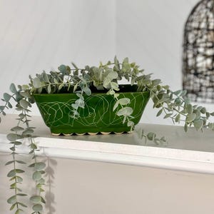 May include: A green ceramic planter, rectangular in shape, with a scalloped base and white abstract pattern. It contains faux eucalyptus greenery. The planter sits on a white shelf, creating a decorative display.