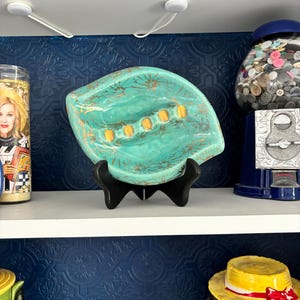 May include: A turquoise ceramic ashtray with gold detailing, presented on a black stand. The ashtray is leaf-shaped, featuring six orange cigarette rests. A gumball machine and a decorative candle are visible in the background.