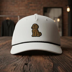 May include: White baseball cap with a brown embroidered poodle design. The cap features a black rope detail along the brim. The hat is displayed on a wooden surface.