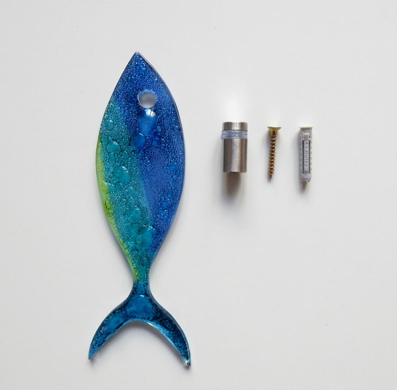 Pu&ograve; includere: Un pesce di vetro blu e verde con una texture a bolle. Il pesce &egrave; appeso a un gancio d'argento e ha un piccolo foro nella parte superiore. Il pesce &egrave; su uno sfondo bianco.