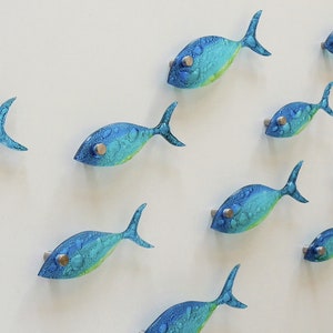 Pu&ograve; includere: Un'opera d'arte da parete con uno scuola di pesci di vetro blu e verde. I pesci sono disposti in uno schema fluido, creando un senso di movimento.
