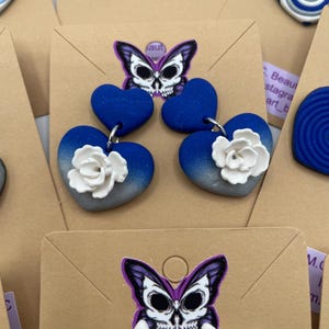 May include: Handmade polymer clay earrings in various designs. One pair features blue heart shapes with white flower accents. Another pair has blue and white spiral designs. Each pair is attached to a brown card with a butterfly skull logo.
