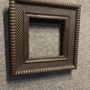 May include: A dark brown, square picture frame with a detailed, textured pattern. The frame has a layered design with a central opening for a photo or artwork. The frame is made of wood and has a classic, vintage aesthetic.