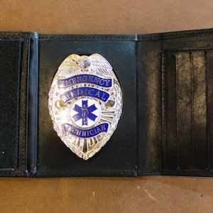 May include: Black leather wallet with an open design, featuring a silver and blue Emergency Medical Technician badge. The badge has a star of life emblem and the words "Emergency Medical Technician". The wallet has multiple card slots.