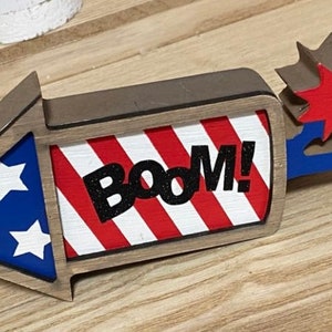 May include: Wooden firework decoration with red, white, and blue stripes and stars. The firework has a brown frame and the word "BOOM!" in black letters.