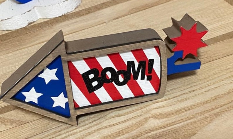 May include: Wooden firework decoration with red, white, and blue stripes and stars. The firework has a brown frame and the word "BOOM!" in black letters.