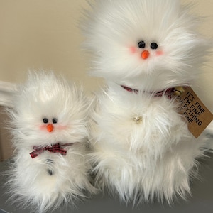 May include: Two fluffy white snowman plush toys. The larger snowman has a red and black plaid scarf, a small bell, and a tag that reads "It's fine, I'm fine, everything's fine." The smaller snowman has a red and black plaid bow tie.