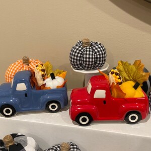 May include: Two ceramic trucks, one blue and one red, are filled with fall-themed decorations. The trucks have white-rimmed wheels and are decorated with pumpkins, corn, and fall leaves. A black and white checkered pumpkin sits on a white pedestal.