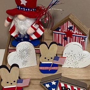 May include: Patriotic themed home decor with a red, white, and blue gnome, two wooden bunny figurines, a wooden heart with fireworks, a wooden house, and a wooden rocket with the word "BOOM!" on it.