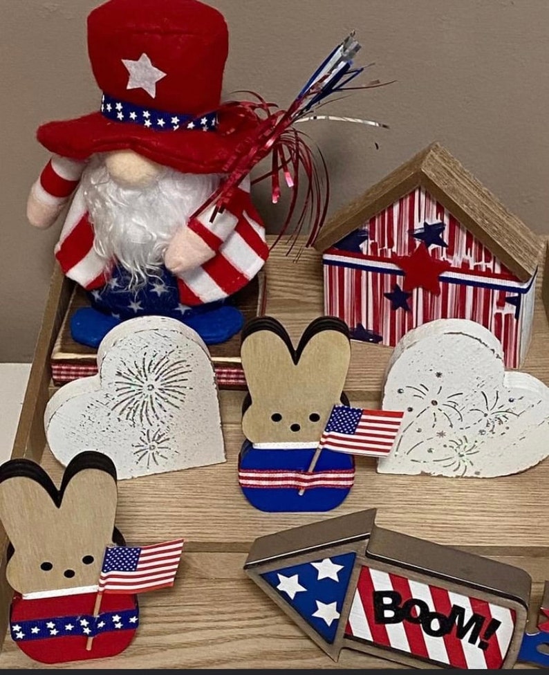May include: Patriotic themed home decor with a red, white, and blue gnome, two wooden bunny figurines, a wooden heart with fireworks, a wooden house, and a wooden rocket with the word "BOOM!" on it.