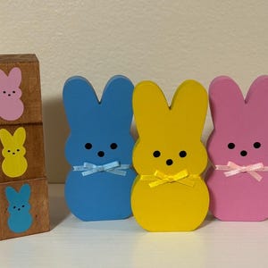 May include: Wooden Easter bunny decorations in blue, yellow, and pink, each with a bow. A stack of three wooden blocks features bunny designs in pink, yellow, and blue. The bunnies have black eyes and are a festive Easter theme.