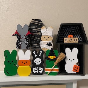 May include: A set of seven Halloween-themed wooden bunny figurines. The bunnies are painted in various colors, including green, yellow, black, white, and gray. They are dressed in costumes, such as a vampire, a mummy, a skeleton, a witch, and a candy corn. The bunnies are arranged on a white shelf with a black wooden house behind them. The house has a black roof and a black and white patterned front. The house has a sign that says "BOO" on it.