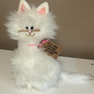 May include: A fluffy white cat plush toy with pink accents on the ears and a pink collar. The cat has a jute twine mustache and a tag that reads "IT'S FINE I'M FINE EVERYTHING IS FINE."