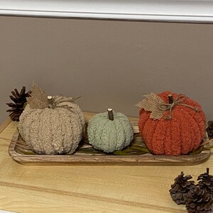 May include: Three knitted pumpkins in a wooden tray. The pumpkins are beige, green, and orange. The tray is decorated with dried grass.