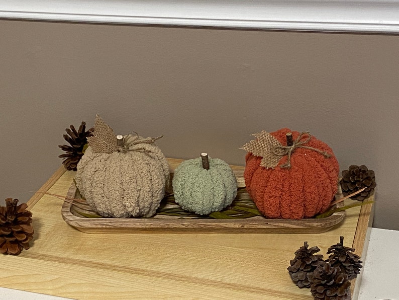 May include: Three knitted pumpkins in a wooden tray. The pumpkins are beige, green, and orange. The tray is decorated with dried grass.