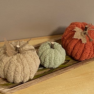 May include: Three knitted pumpkins in different sizes and colors: beige, green, and orange. The pumpkins are arranged on a wooden tray with green leaves.