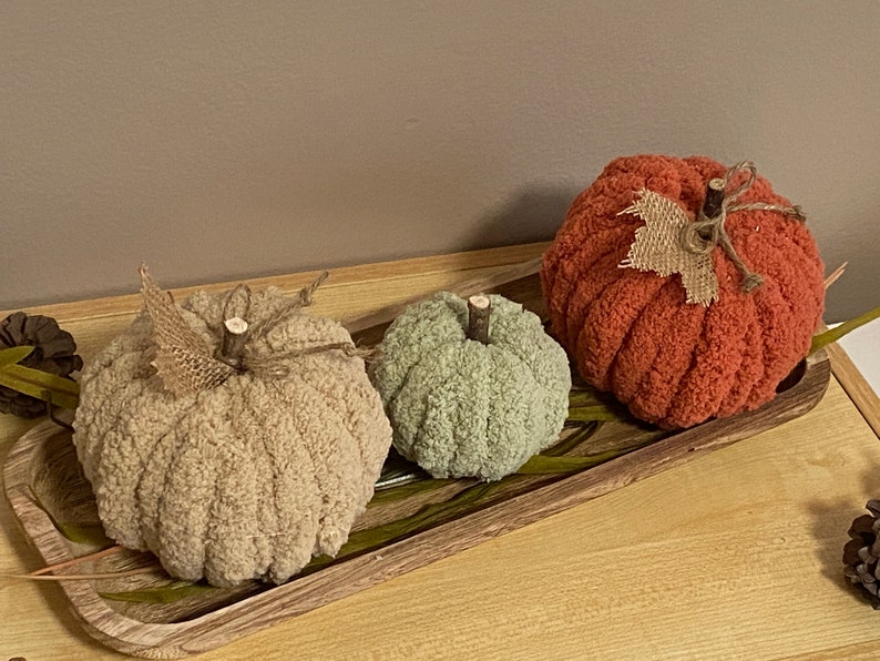 May include: Three knitted pumpkins in different sizes and colors: beige, green, and orange. The pumpkins are arranged on a wooden tray with green leaves.