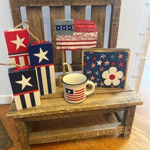 May include: A rustic wooden bench displays patriotic-themed decor. Items include red, white, and blue star-adorned blocks, a miniature flag, a small mug with a flag design, and a framed floral design. The items are arranged on a wooden bench.