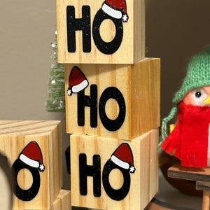 May include: Wooden blocks stacked to spell "HO HO HO" with a red Santa hat on each block. The blocks are a light brown color and the letters are black.