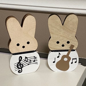May include: Two wooden bunny figurines, one with a musical note design and the other with a guitar design. Both bunnies are painted in a light brown color and have black eyes.