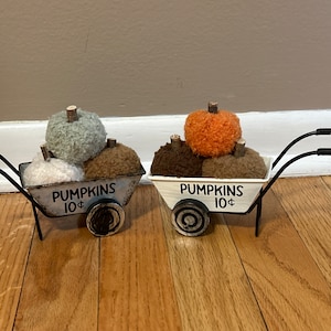 May include: Two miniature wheelbarrows filled with fuzzy pumpkins. Each wheelbarrow is painted white with black lettering that reads "Pumpkins 10¢".