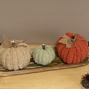 May include: Three knitted pumpkins in a wooden tray. The pumpkins are beige, green, and orange. The pumpkins have burlap leaves and twine accents.