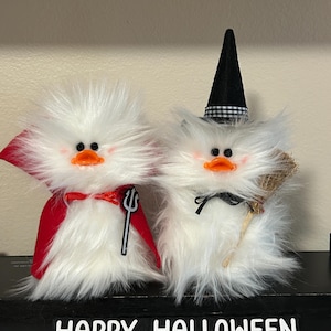 May include: Two fluffy white Halloween decorations. One is dressed as a devil with a red cape and trident. The other is a witch with a black hat and broom. The words "HAPPY HALLOWEEN" are displayed below.