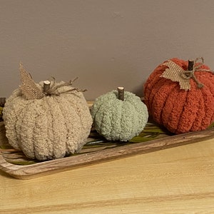 May include: Three knitted pumpkins in a wooden tray. The pumpkins are beige, green, and orange. The tray is brown and has a wood grain pattern.