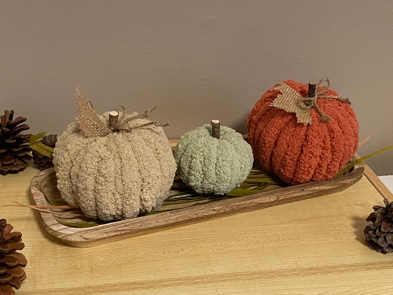 May include: Three knitted pumpkins in a wooden tray. The pumpkins are beige, green, and orange. The tray is brown and has a wood grain pattern.