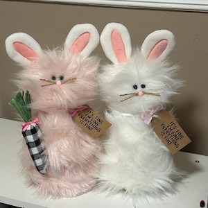 May include: Two plush bunny rabbits, one pink and one white, with embroidered faces and long ears. Each bunny holds a sign that reads "It's fine, I'm fine, everything is fine."
