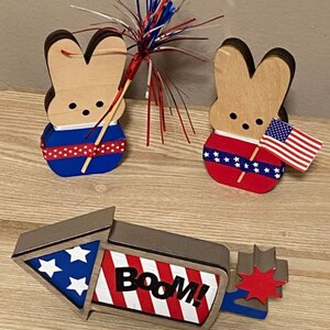 May include: Three wooden patriotic decorations for the Fourth of July. Two are bunny shaped with red, white, and blue decorations. One is a rocket shaped with red, white, and blue stripes and the word "BOOM!" in black.