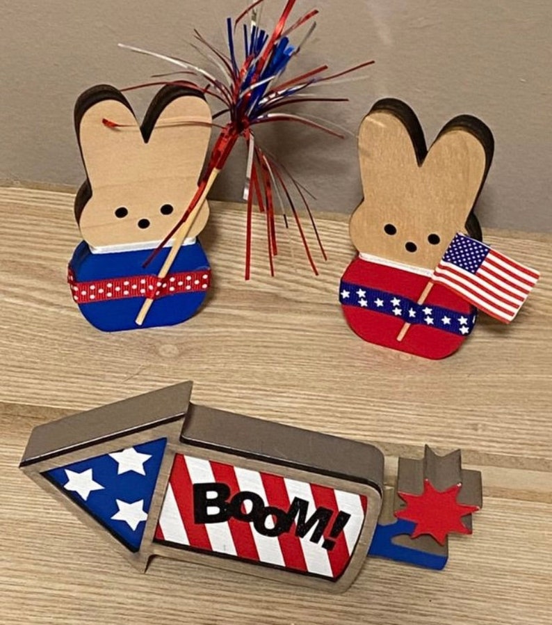 May include: Three wooden patriotic decorations for the Fourth of July. Two are bunny shaped with red, white, and blue decorations. One is a rocket shaped with red, white, and blue stripes and the word "BOOM!" in black.