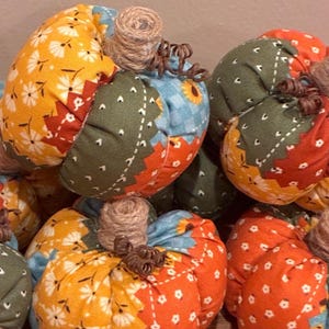 May include: A collection of stuffed fabric pumpkins featuring patchwork designs in orange, green, and blue floral patterns. Each pumpkin is topped with a wrapped jute twine stem and a small, curled brown wire accent, creating a textured appearance.