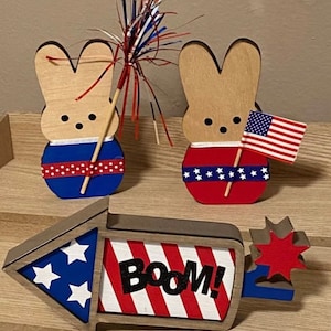 May include: Two wooden bunny figurines decorated in red, white, and blue with a red, white, and blue firework and a wooden sign that says "Boom!" in black letters.