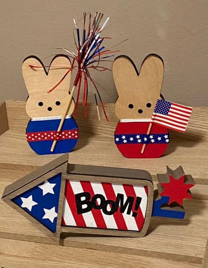 May include: Two wooden bunny figurines decorated in red, white, and blue with a red, white, and blue firework and a wooden sign that says "Boom!" in black letters.