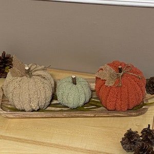 May include: Three knitted pumpkins in a wooden tray. The pumpkins are beige, green, and orange. The tray has a wood grain pattern and is decorated with dried leaves.