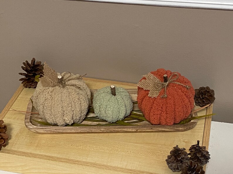 May include: Three knitted pumpkins in a wooden tray. The pumpkins are beige, green, and orange. The tray has a wood grain pattern and is decorated with dried leaves.