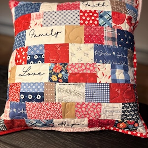 May include: A patchwork pillow with a red, white, and blue color scheme. The pillow features the words "Family," "Love," "Faith," and "Hope" embroidered on it. The pillow also has a recipe for Grandma's Lemon Meringue Pie.