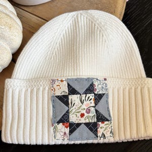 May include: A cream-colored ribbed knit beanie featuring a square fabric patch on the front. The patch displays a geometric star pattern composed of dark gray, light blue, and floral-printed fabric squares stitched together.
