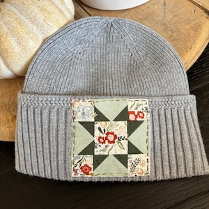 May include: A light gray ribbed knit beanie featuring a square fabric patch on the front cuff. The patch displays a star-shaped quilt pattern in shades of sage green, white, and floral prints with red and orange flowers, secured with visible stitching.