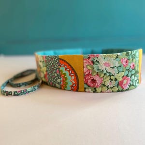 Puede incluir: Un cinturón de tela colorido con un estampado floral y un estampado geométrico amarillo, naranja y verde. El cinturón está atado con un cordón de tela a juego.