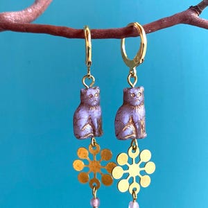 Op de afbeelding: Een paar goudkleurige oorringen. Elke oorring heeft een lavendelkleurige kattenbedel met gouden accenten, een gouden sneeuwvlokbedel en een kleine roze kraal. De achtergrond is helderblauw.