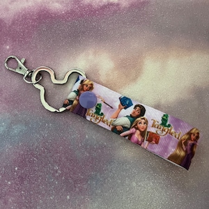 May include: A purple and white key chain with a Mickey Mouse shaped metal ring. The key chain features a printed image of Rapunzel and Flynn Rider from the Disney movie Tangled. The text "Tangled" is printed on the key chain.