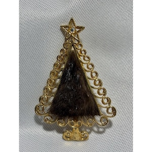 May include: A gold-toned Christmas tree brooch with a star on top. The tree's center is filled with brown fur, and the edges are adorned with decorative swirls. A small, clear gem is set in the star.