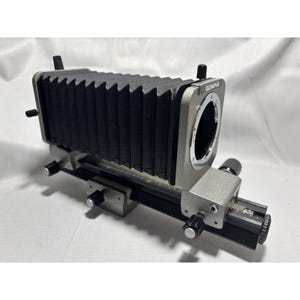 May include: An Olympus bellows macro focusing system. The device features a black bellows, a silver-colored body, and multiple adjustment knobs. The Olympus logo is visible on the side. This system is designed for macro photography.