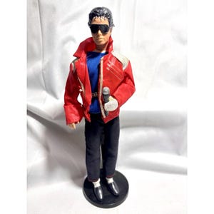 May include: A doll of a male figure wearing a red jacket with white accents, a blue shirt, and black pants. The doll is holding a microphone and wearing sunglasses. The doll is standing on a black base.